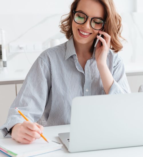 Portrait of beautiful smiling woman in glasses taking notes while speaking on mobile phone, indoors, pannels solar